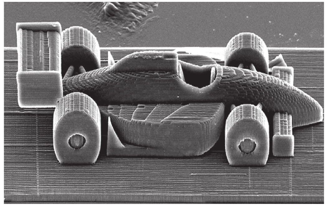 Fotografia de microscopia eletrônica, em tons de cinza, mostrando a miniatura de um carro de Fórmula 1 esculpida sobre uma superfície estriada. Vê-se o carro de perfil, com as quatro rodas, o bico alongado à direita, o cockpit no meio e as asas dianteira e traseira; a textura em camadas da impressão aparece em toda a peça.