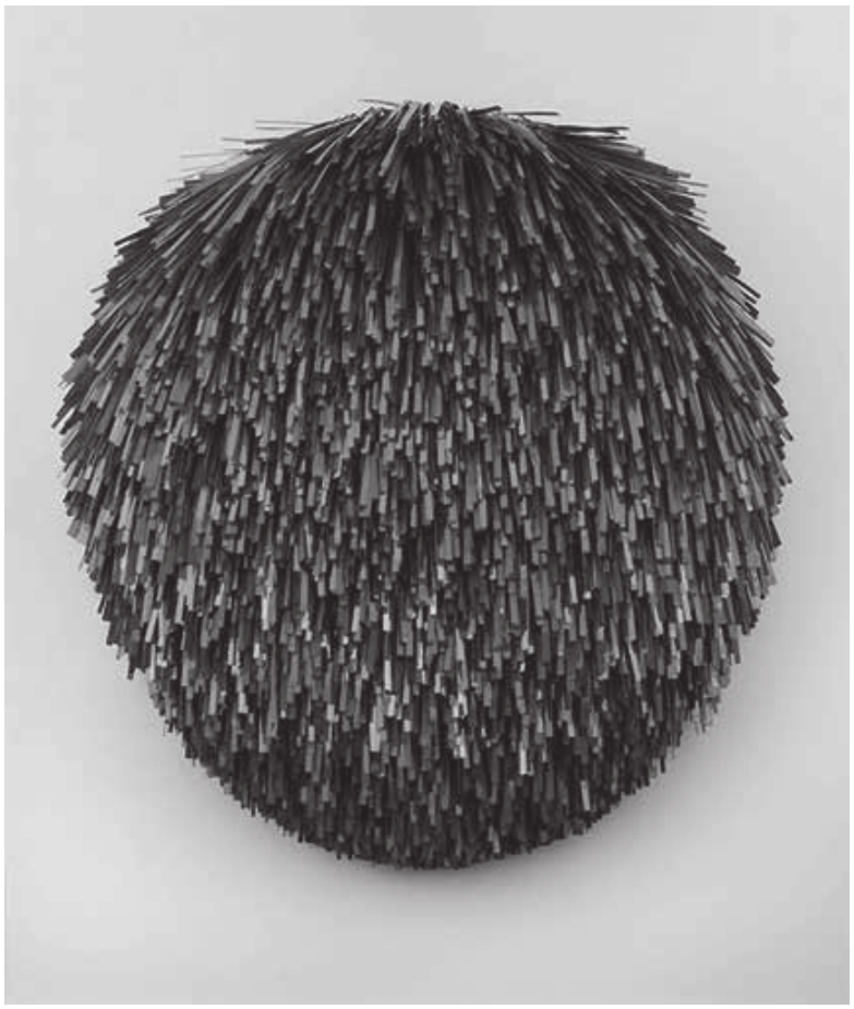 Fotografia em preto e branco de uma escultura de parede, quase esférica e achatada embaixo, formada por milhares de hastes finas e escuras de metal apontadas para fora, mais densas no centro e eriçadas nas bordas. O fundo é claro e liso, e a peça projeta sombra suave sobre ele.
