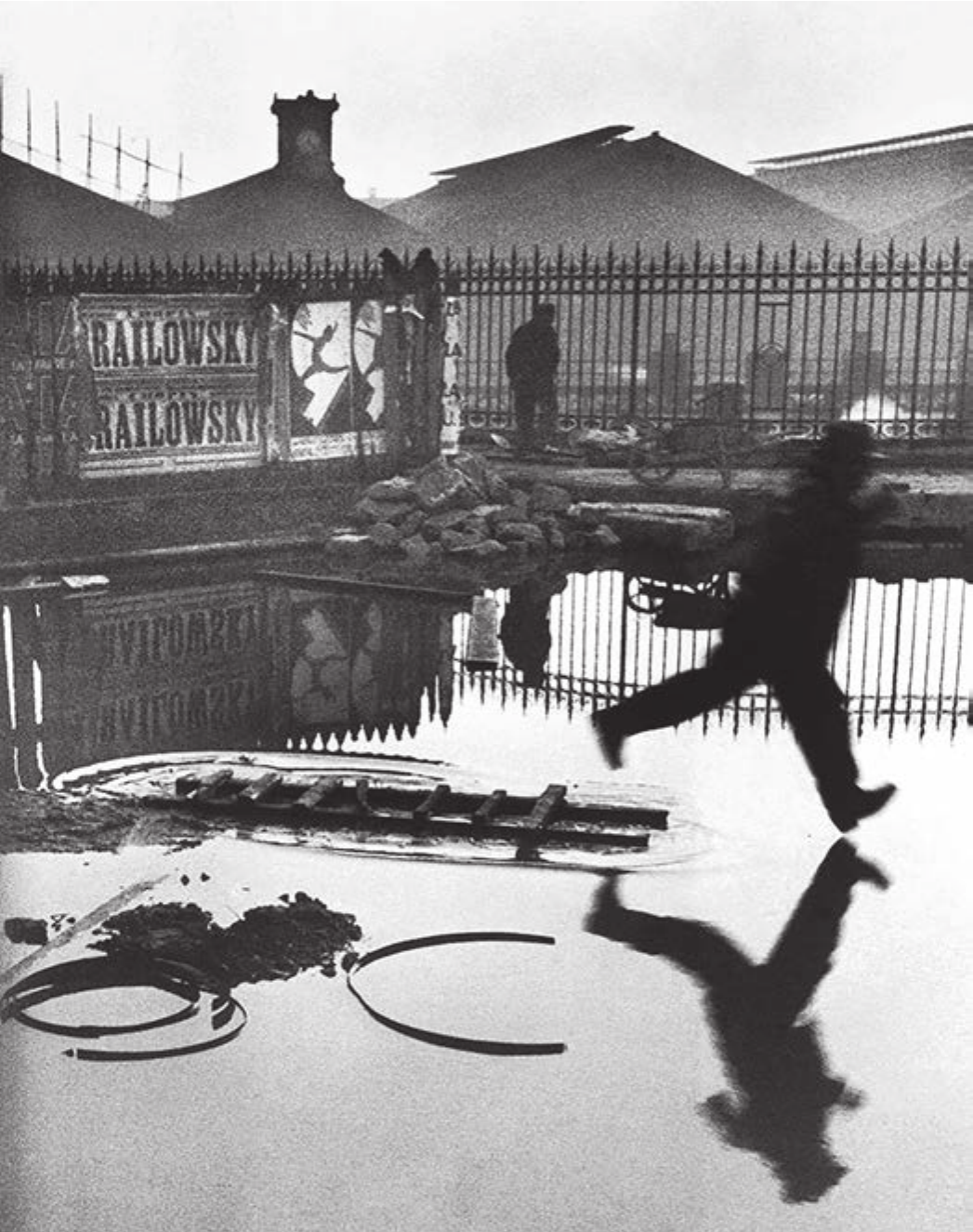 Fotografia em preto e branco de Henri Cartier-Bresson, “Atrás da estação de Saint-Lazare”. Um homem de chapéu salta sobre uma grande poça de água, com o corpo suspenso no ar e o calcanhar quase tocando a superfície; seu reflexo aparece invertido na água, em simetria quase perfeita. Ao fundo, uma cerca de ferro, telhados de estação, escadas e entulho; à esquerda, cartazes com a inscrição repetida “RAILOWSKY”, também refletidos de cabeça para baixo. No primeiro plano, aros metálicos e uma escada deitada sobre a água.