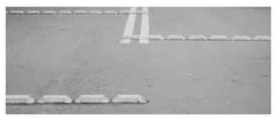 Fotografia em preto e branco de um trecho de pista asfaltada com fileiras de tachões: peças claras, baixas e alongadas, com as laterais inclinadas, fixadas no chão uma após a outra, formando linhas que delimitam faixas.
