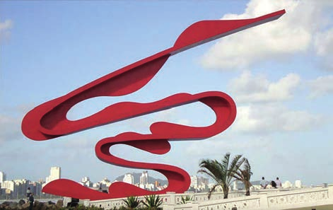 Fotografia de uma escultura metálica vermelha, de faixas largas curvadas em laços contínuos, instalada ao ar livre sobre um terraço com balaustrada branca e palmeiras; ao fundo, o céu com nuvens e a silhueta de prédios da cidade.