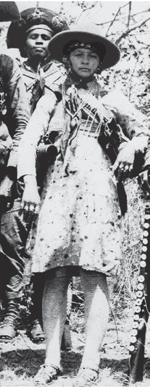 Fotografia em preto e branco, granulada, dos anos 1930: Maria Bonita, cangaceira, em pé, de chapéu de abas largas com enfeites, lenço no pescoço, vestido claro estampado até os joelhos, cartucheira cruzada e cordões; atrás dela, um cangaceiro de chapéu de couro, entre a vegetação da caatinga.