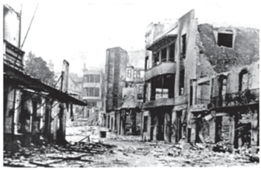 Fotografia em preto e branco de uma rua em ruínas: prédios com fachadas destruídas, janelas vazias, vigas e paredes desabadas, entulho espalhado pelo chão; não há pessoas na cena.