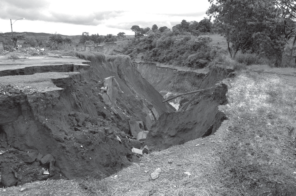 Fotografia em preto e branco de uma voçoroca em área urbana: uma vala profunda e larga, de paredes de terra desmoronadas, corta um terreno aberto com capim e entulho; ao fundo aparecem árvores, um poste de iluminação e construções.