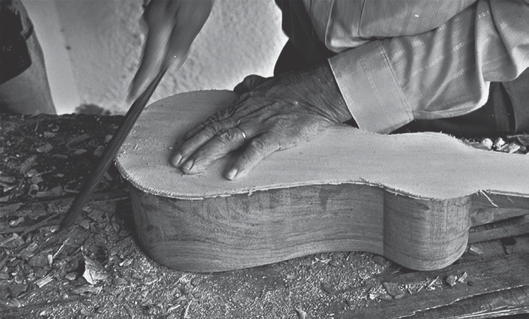 Fotografia em preto e branco, em primeiro plano: as mãos de um artesão apoiando e entalhando com um formão o tampo de madeira de um instrumento de cordas (viola) em construção; sobre a bancada, lascas e serragem.