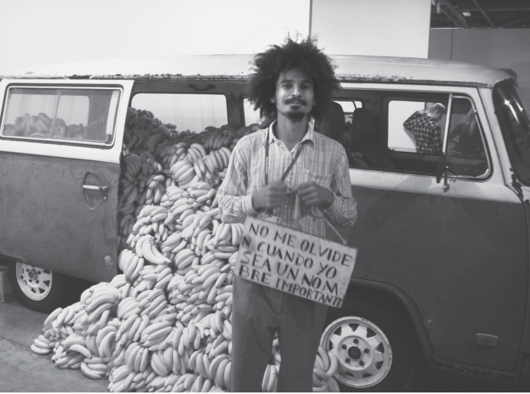 Fotografia em preto e branco: uma Kombi com a porta lateral aberta, lotada de cachos de bananas que se derramam para fora; à frente, um homem negro de cabelo volumoso, camisa clara, segura uma placa de papelão com a inscrição em espanhol “NO ME OLVIDE CUANDO YO SEA UN NOMBRE IMPORTANTE”. Ao fundo, um ambiente interno de feira.
