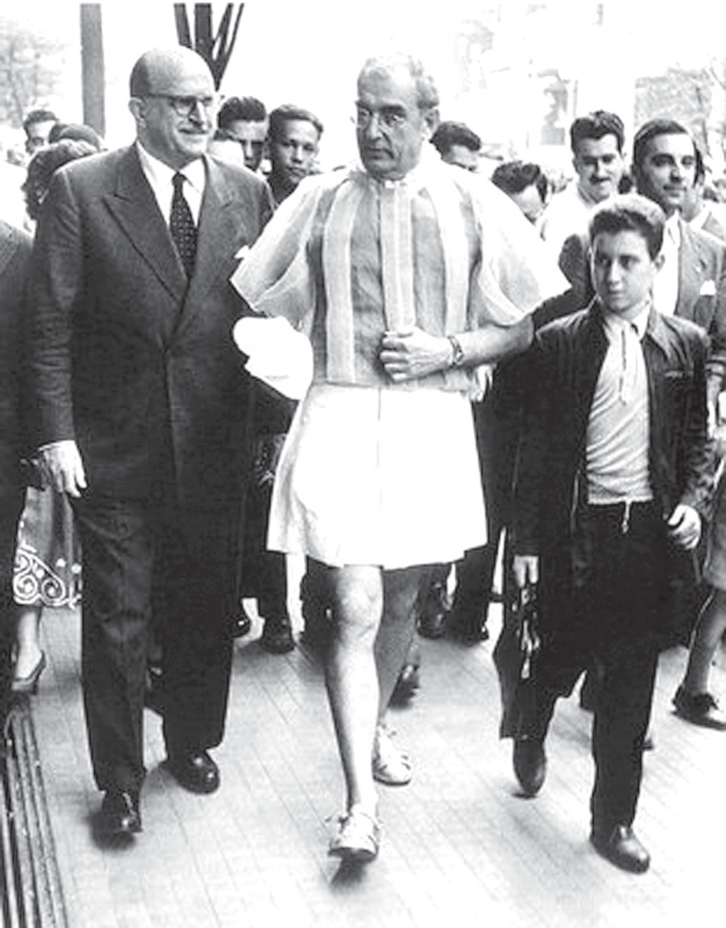 Fotografia em preto e branco, de 1956, de um homem calvo e de óculos caminhando por uma calçada de avenida, cercado por uma multidão. Ele veste uma blusa listrada de mangas bufantes e uma saia curta clara, com meias e sapatos baixos, deixando as pernas à mostra. Ao seu lado, um homem de terno e gravata e um rapaz de jaqueta; ao fundo, várias pessoas observam e o seguem.
