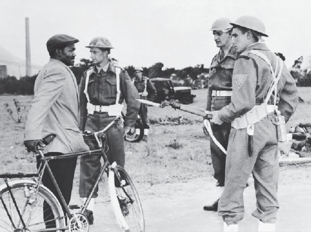 Fotografia em preto e branco: um homem negro, de boné e paletó, segurando uma bicicleta, é abordado em uma estrada por três soldados brancos de capacete e uniforme, um deles apontando um fuzil com baioneta; ao fundo, um veículo militar e chaminés.