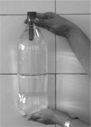 Fotografia de uma garrafa PET transparente cheia de água, segurada por uma mão; dentro dela, junto ao gargalo, na parte superior, flutua um pequeno frasco escuro emborcado.
