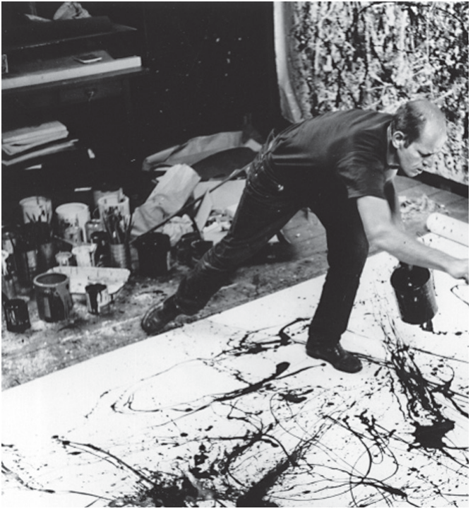 Fotografia em preto e branco de um ateliê. Um homem calvo, de camiseta e calça escuras, inclina-se sobre uma grande tela estendida no chão e, com uma lata de tinta na mão, deixa a tinta escorrer e respingar sobre ela, formando linhas e manchas emaranhadas. Ao fundo, muitas latas e potes de tinta com pincéis e outra tela coberta de respingos encostada na parede.