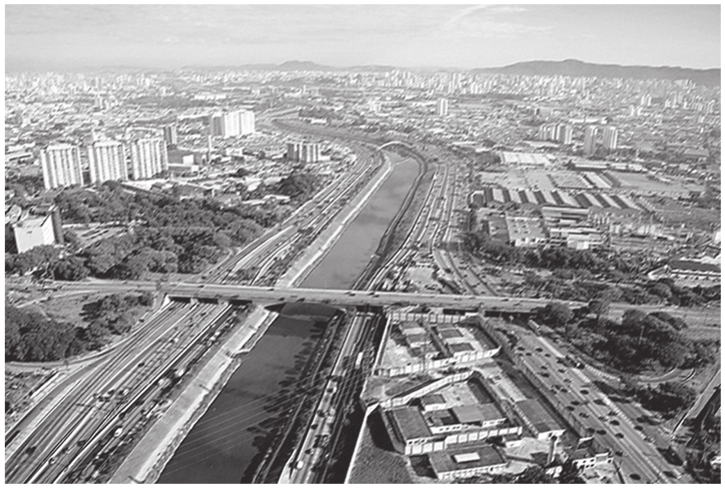 Fotografia aérea de uma cidade cortada por um rio retificado entre marginais movimentadas: de um lado, prédios altos e árvores; do outro, galpões e pátios industriais; ao fundo, a mancha urbana até o horizonte. Uma ponte larga atravessa o rio no primeiro plano, e a água aparece escura e opaca.