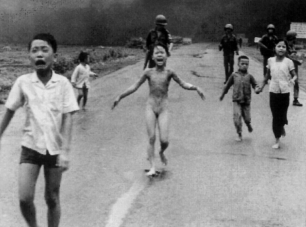 Fotografia em preto e branco de uma estrada de terra: em primeiro plano, crianças correm em direção ao observador com expressões de pavor; um menino de camisa clara grita, e no centro uma menina nua, com os braços abertos, chora; outras crianças correm ao lado, uma delas de mãos dadas com uma mais velha. Ao fundo, soldados com capacete e armas caminham pela estrada e o céu está tomado por fumaça escura.